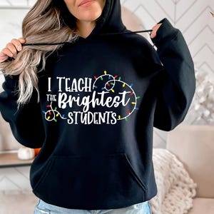 May include: Black hooded sweatshirt with the text "I Teach the Brightest Students" in white, surrounded by a string of colorful Christmas lights. The sweatshirt has a front pocket and a drawstring hood.