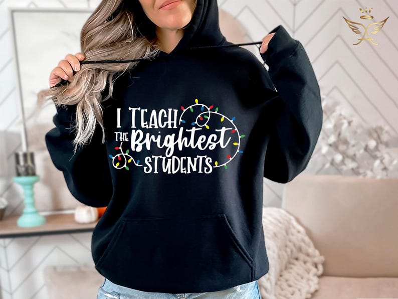 May include: Black hooded sweatshirt with the text "I Teach the Brightest Students" in white, surrounded by a string of colorful Christmas lights. The sweatshirt has a front pocket and a drawstring hood.