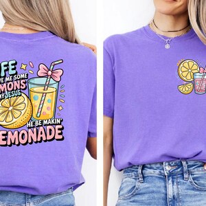 May include: Purple t-shirts with lemonade-themed graphics. One shirt features a large graphic on the back with text and a lemonade glass. The other shirt has a smaller lemonade graphic on the front. Both shirts have a crew neck.