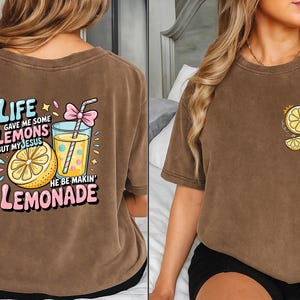 May include: Brown t-shirt with a colorful graphic of a lemon, lemonade, and text that reads "Life gave me some lemons but my Jesus he be makin' lemonade." A smaller version of the graphic is on the front.