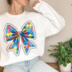Puede incluir: Sudadera blanca con un dise&ntilde;o de lazo de piezas de rompecabezas azules, rojas y amarillas. El texto alrededor del lazo dice "No te cambiar&iacute;a por el mundo, pero cambiar&iacute;a el mundo por ti".