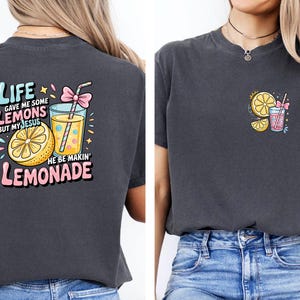 May include: Dark gray t-shirt with a colorful graphic on the back that says "Life gave me some lemons but my Jesus he be makin' lemonade." The design includes a lemon, a glass of lemonade, and a pink bow. A smaller version of the design is on the front.