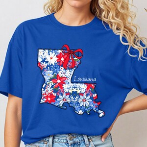 May include: A royal blue t-shirt with a floral design of the state of Louisiana. The design includes a red bow and the word "Louisiana".