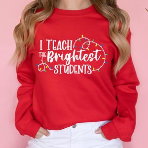 May include: A red sweatshirt with the text "I Teach the Brightest Students" in white, surrounded by a string of colorful Christmas lights. The sweatshirt has long sleeves and is worn with white pants.