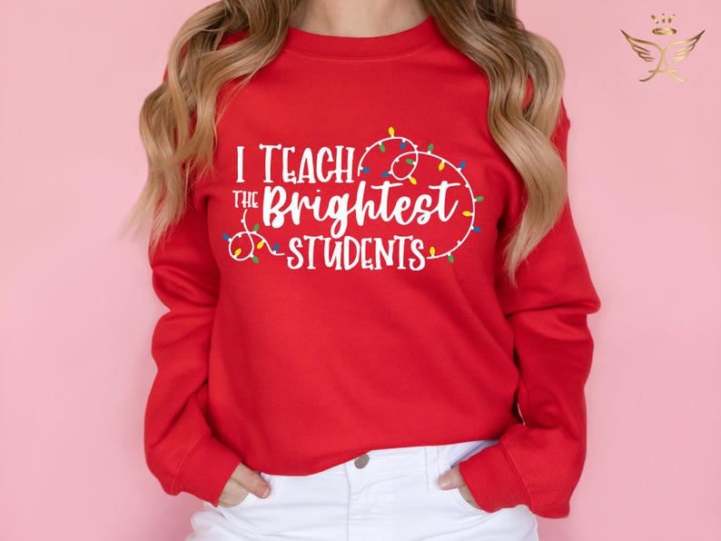 May include: A red sweatshirt with the text "I Teach the Brightest Students" in white, surrounded by a string of colorful Christmas lights. The sweatshirt has long sleeves and is worn with white pants.
