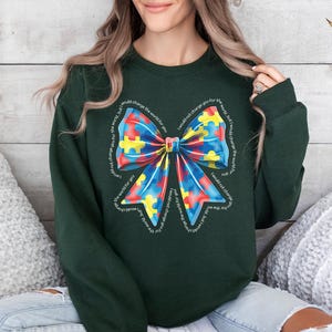 Puede incluir: Una sudadera verde oscuro con un gran lazo colorido hecho de piezas de rompecabezas. El lazo est&aacute; rodeado por el texto "No te cambiar&iacute;a por el mundo, pero cambiar&iacute;a el mundo por ti".