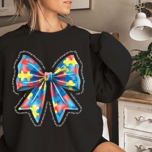 Puede incluir: Sudadera negra con un lazo de piezas de rompecabezas de colores y el texto "No te cambiar&iacute;a por el mundo, pero cambiar&iacute;a el mundo por ti".