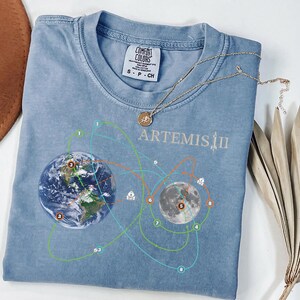 May include: A light blue Comfort Colors t-shirt with a graphic of Earth and the Moon, connected by lines, and the text "ARTEMIS II". A gold necklace with a pendant rests on the shirt. A brown hat and dried leaves are in the background.