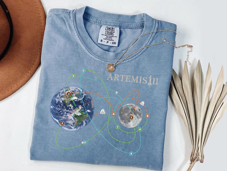 May include: A light blue Comfort Colors t-shirt with a graphic of Earth and the Moon, connected by lines, and the text "ARTEMIS II". A gold necklace with a pendant rests on the shirt. A brown hat and dried leaves are in the background.