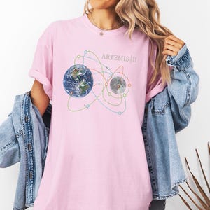 May include: A light pink t-shirt featuring a graphic of Earth and the Moon with orbital paths, and the text "ARTEMIS III". The design includes blue, green, and yellow lines. The shirt is a soft, comfortable material.