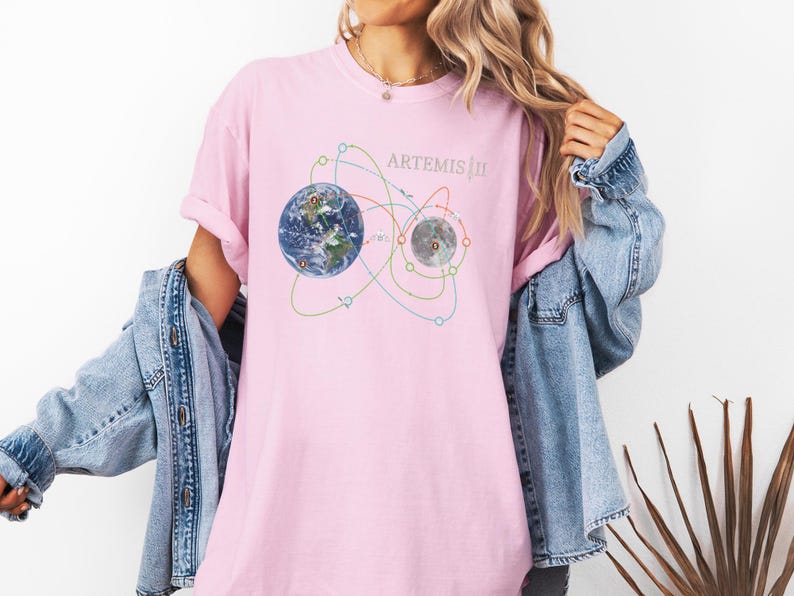 May include: A light pink t-shirt featuring a graphic of Earth and the Moon with orbital paths, and the text "ARTEMIS III". The design includes blue, green, and yellow lines. The shirt is a soft, comfortable material.