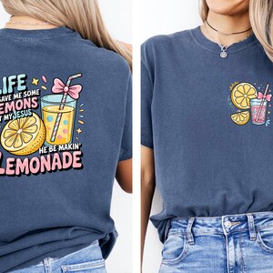 May include: Navy blue t-shirt with a colorful graphic of a lemon, lemonade, and text that reads "Life gave me some lemons but my Jesus he be makin' lemonade." A smaller version of the graphic is on the front.