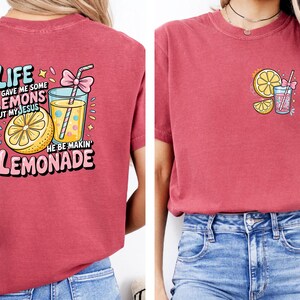May include: Red t-shirt with a colorful graphic on the front and back. The front graphic features a lemon slice and a drink with a straw. The back graphic says "Life gave me some lemons but my Jesus he be makin' lemonade."