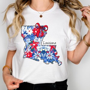 May include: White t-shirt with a floral print of the state of Louisiana. The print includes a red bow and the word "Louisiana".
