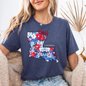 May include: A navy blue t-shirt with a floral print of the state of Louisiana. The print includes a red bow and the word "Louisiana".