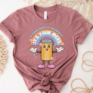 Peut inclure: Un t-shirt mauve avec un arc-en-ciel et une pilule de dessin anim&eacute; avec des bras et des jambes. Le texte sur le t-shirt est "Si tu es heureux et que tu le sais, c'est tes m&eacute;dicaments."