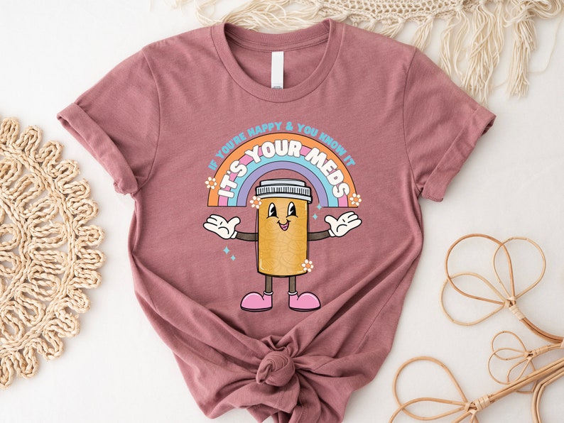 Peut inclure: Un t-shirt mauve avec un arc-en-ciel et une pilule de dessin anim&eacute; avec des bras et des jambes. Le texte sur le t-shirt est "Si tu es heureux et que tu le sais, c'est tes m&eacute;dicaments."