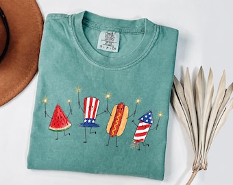 May include: A sage green t-shirt featuring a graphic print of a smiling watermelon slice, a top hat with stars and stripes, a hot dog, and a firework, all holding sparklers. The inner neck label reads Comfort Colors, 100% cotton, size S, P, CH.
