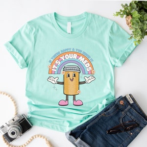 Peut inclure: Un t-shirt bleu clair avec un arc-en-ciel et une pilule de dessin anim&eacute; avec un visage souriant. Le texte sur le t-shirt est "Si vous &ecirc;tes heureux et que vous le savez, c'est vos m&eacute;dicaments".