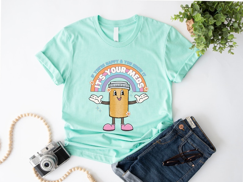 Peut inclure: Un t-shirt bleu clair avec un arc-en-ciel et une pilule de dessin anim&eacute; avec un visage souriant. Le texte sur le t-shirt est "Si vous &ecirc;tes heureux et que vous le savez, c'est vos m&eacute;dicaments".