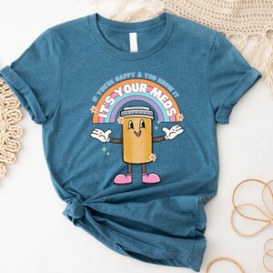 Peut inclure: Un t-shirt bleu sarcelle avec un personnage de pilule de dessin anim&eacute; et un arc-en-ciel. Le texte sur le t-shirt est "If you're happy & you know it, it's your meds".