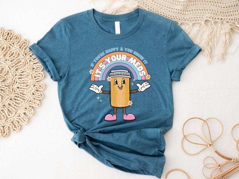 Peut inclure: Un t-shirt bleu sarcelle avec un personnage de pilule de dessin anim&eacute; et un arc-en-ciel. Le texte sur le t-shirt est "If you're happy & you know it, it's your meds".