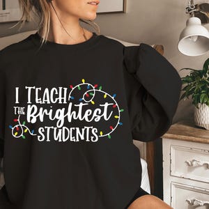 May include: Black sweatshirt with the text "I Teach the Brightest Students" in white, accented with a string of colorful Christmas lights. The sweatshirt is a crew neck style.