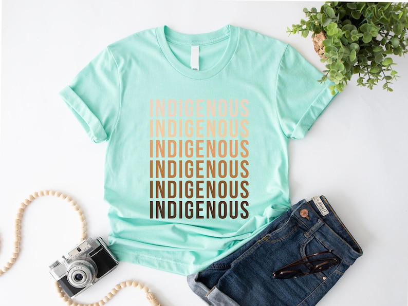 Może przedstawiać: Jasno błękitny t-shirt z napisem "Indigenous" powt&oacute;rzonym sześć razy w gradiencie brązu, pomarańczy i beżu.