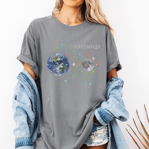 May include: Gray t-shirt featuring a graphic of Earth and the Moon connected by colorful lines, with the text "ARTEMIS II" in white. The design suggests a space exploration theme. The shirt is worn with a denim jacket and shorts.
