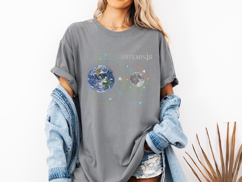 May include: Gray t-shirt featuring a graphic of Earth and the Moon connected by colorful lines, with the text "ARTEMIS II" in white. The design suggests a space exploration theme. The shirt is worn with a denim jacket and shorts.