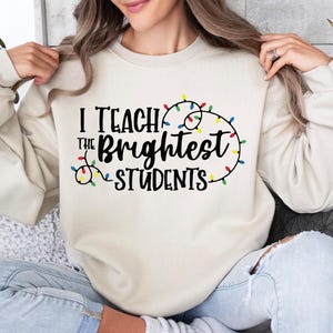 May include: Cream-colored sweatshirt with the text "I Teach the Brightest Students" in black, accented with a string of colorful Christmas lights. The long-sleeved top is a casual style.