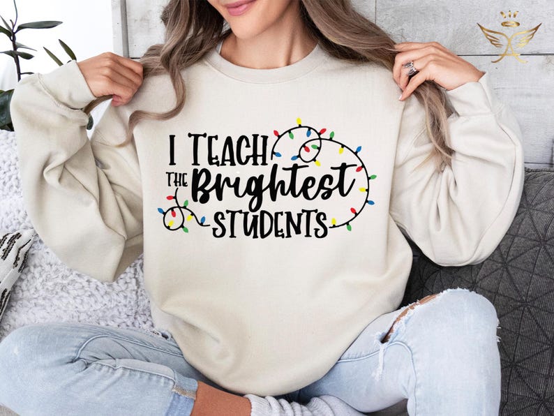 May include: Cream-colored sweatshirt with the text "I Teach the Brightest Students" in black, accented with a string of colorful Christmas lights. The long-sleeved top is a casual style.