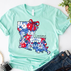 May include: A light blue t-shirt with a floral print of the state of Louisiana. The print includes a red bow and the word "Louisiana".