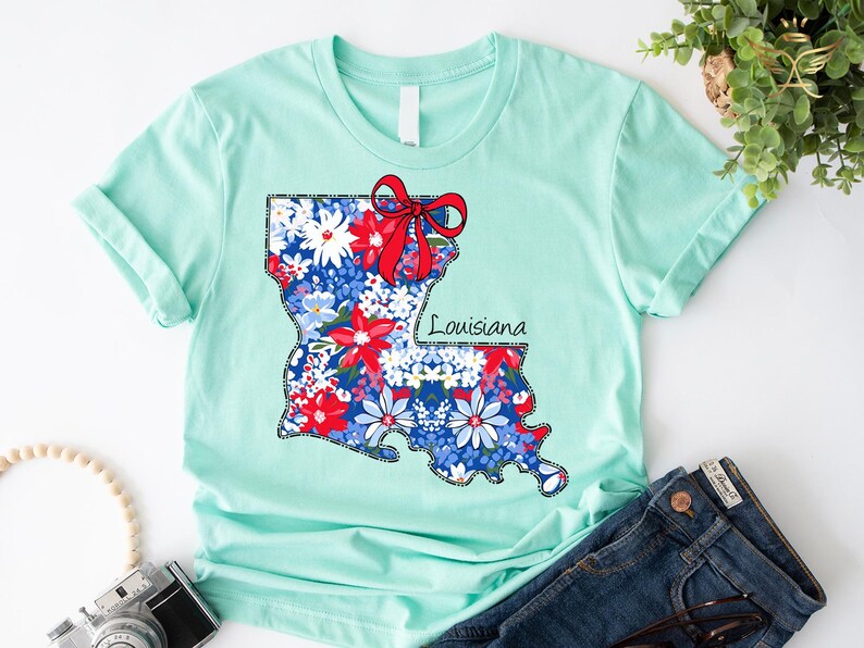 May include: A light blue t-shirt with a floral print of the state of Louisiana. The print includes a red bow and the word "Louisiana".