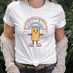 Peut inclure: T-shirt blanc avec un arc-en-ciel et une pilule de dessin anim&eacute; avec le texte "Si vous &ecirc;tes heureux et que vous le savez, c'est vos m&eacute;dicaments".