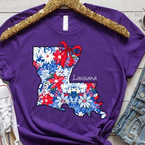 May include: Purple t-shirt with a floral design of the state of Louisiana with a red bow. The text "Louisiana" is printed on the shirt.