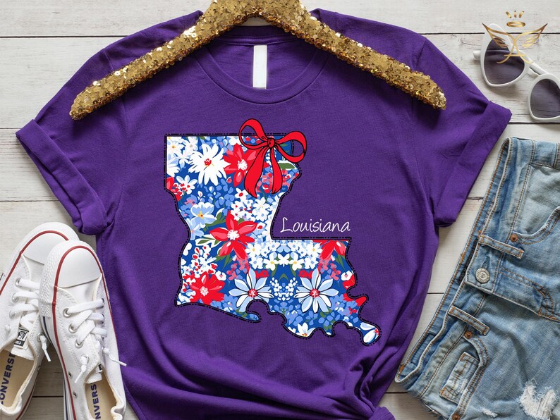 May include: Purple t-shirt with a floral design of the state of Louisiana with a red bow. The text "Louisiana" is printed on the shirt.