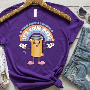 Peut inclure: T-shirt violet avec un arc-en-ciel et une pilule de dessin anim&eacute; avec le texte "Si vous &ecirc;tes heureux et que vous le savez, c'est vos m&eacute;dicaments".