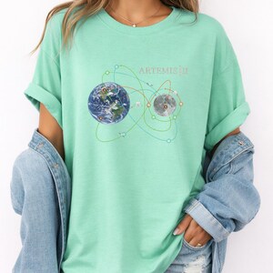 May include: A light green t-shirt featuring a graphic of Earth and the Moon connected by lines, with the text "ARTEMIS III" above. The shirt is worn with a denim jacket.