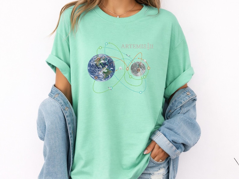 May include: A light green t-shirt featuring a graphic of Earth and the Moon connected by lines, with the text "ARTEMIS III" above. The shirt is worn with a denim jacket.