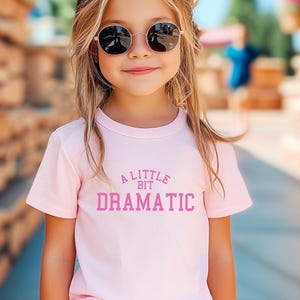 May include: A young girl wearing a pink t-shirt with the text "A Little Bit Dramatic" in pink lettering. She is wearing sunglasses and has blonde hair.
