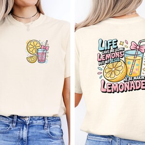 May include: Cream-colored t-shirt with a colorful lemonade-themed graphic. The front features a lemon slice, a drink with a straw, and the text "Life gave me some lemons." The back has the text "Life gave me some lemons but my Jesus he be makin' lemonade."