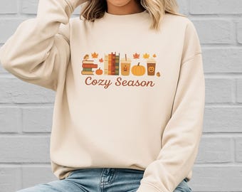 May include: Cream-colored sweatshirt with a fall-themed graphic. The design includes stacks of books, a pumpkin, coffee cups, and autumn leaves. The words "Cozy Season" are written in a brown script font.