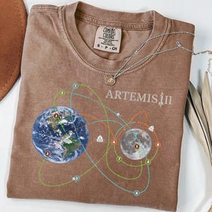 May include: A brown t-shirt with a graphic design featuring Earth, the Moon, and connecting orbital paths. The text "ARTEMIS II" is printed above the moon graphic. A silver necklace with a pendant is draped on the shirt.