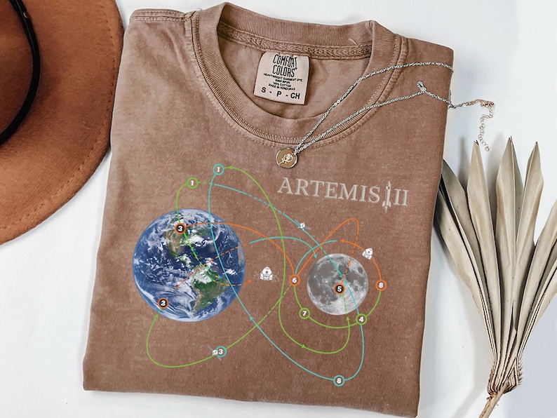 May include: A brown t-shirt with a graphic design featuring Earth, the Moon, and connecting orbital paths. The text "ARTEMIS II" is printed above the moon graphic. A silver necklace with a pendant is draped on the shirt.