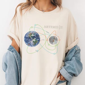 May include: Cream-colored t-shirt featuring a graphic of Earth and the Moon connected by colorful orbital paths. The text "ARTEMIS III" is printed above the graphic. The shirt is worn with a denim jacket.