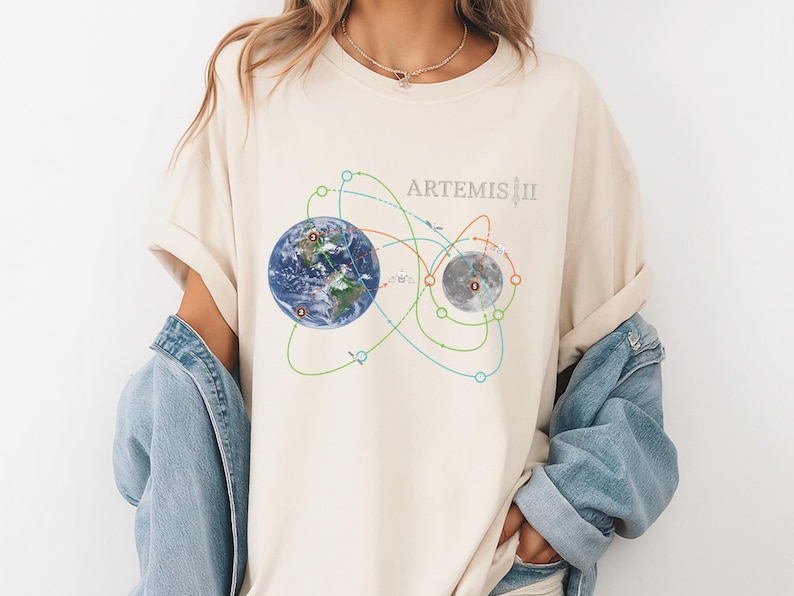 May include: Cream-colored t-shirt featuring a graphic of Earth and the Moon connected by colorful orbital paths. The text "ARTEMIS III" is printed above the graphic. The shirt is worn with a denim jacket.
