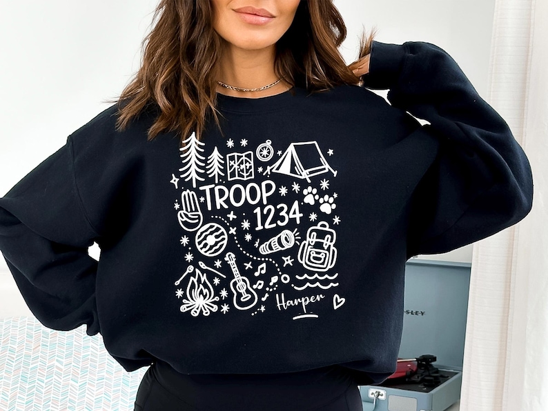 May include: Black sweatshirt with a white graphic design featuring camping-themed illustrations, including trees, a tent, a compass, and a guitar. The text "TROOP 1234" is also included.