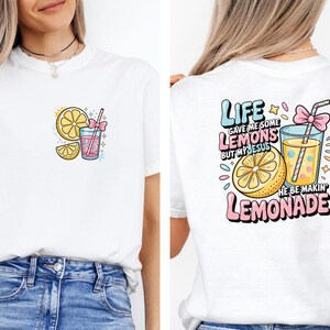 May include: White t-shirt featuring a colorful graphic of a lemon, lemonade, and the text "Life gave me some lemons but my Jesus he be makin' lemonade." The design includes pink, yellow, and blue accents. The shirt is a casual wear item.