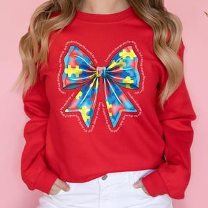 Puede incluir: Sudadera roja con un gran lazo azul, amarillo y rojo hecho de piezas de rompecabezas. El lazo est&aacute; rodeado por el texto "No cambiar&iacute;a el mundo por ti, pero cambiar&iacute;a el mundo por ti".